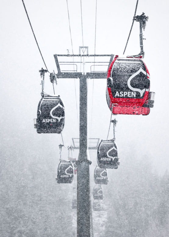 Red and black Aspen Snowmass gondolas move upward in heavy snowfall, surrounded by snowy trees.