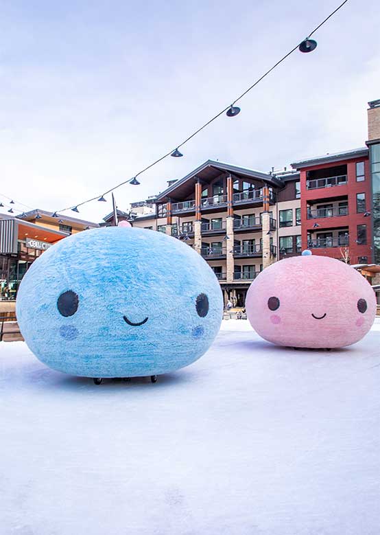 Two large, smiling, plush-like figures, one blue and one pink, sit on a snowy ground.