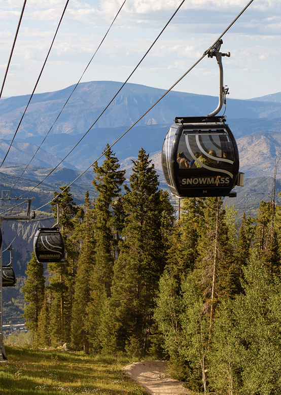 Gondola cars traveling uphill at Snowmass, during the summer. Valley spans behind the black cable cars, trees line the path of the gondola.