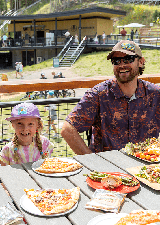 Father and daughter smile at the camera, at Ek camp in the summer. Fresh slices of pizza, veggies, salads and a cookie sit on the table in front of them.