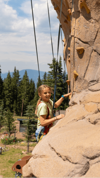 Little girl looks over an edge of rocks while she climbs on the outdoor climbing wall at camp aspen snowmass