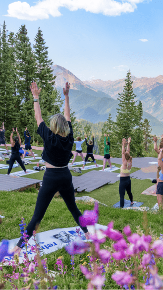 Group of people do yoga atop aspen mountain, surrounded by wildflowers under a blue sky