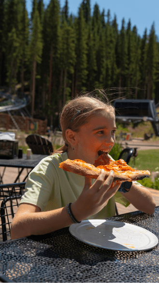 Little girl takes bite of large slice of pizza, sitting outside at Elk Camp Snowmass