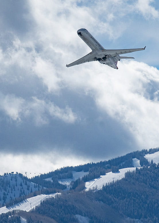 An airplane climbs steeply through the sky, above snow-covered mountains dotted with trees.