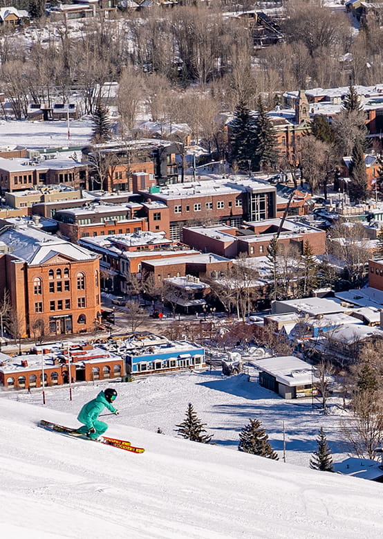 A skier in green swiftly descends a snowy slope, with a snow-dusted town below.
