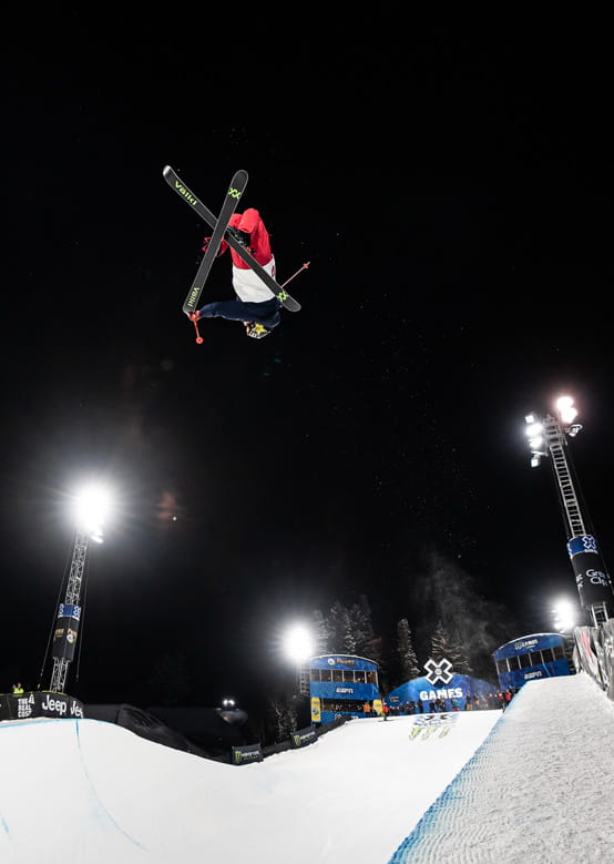 A skier performs a jump with crossed skis at the X Games, surrounded by illuminated structures.