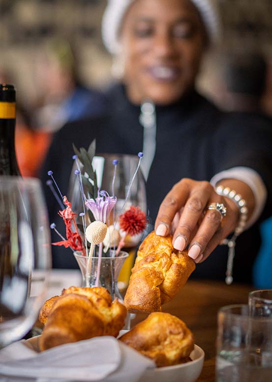 A hand picks up a golden pastry from a bowl surrounded by wine glasses and decorative flowers.
