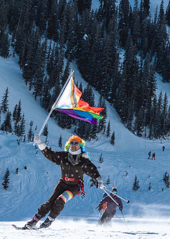 A skier waves a rainbow flag while descending a snowy mountain slope surrounded by trees at Aspen's Gay Ski Week.