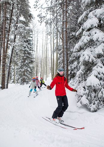 A ski instructor leads kids down a forested trail at Snowmass