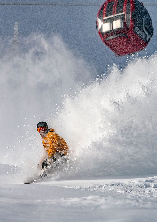 A snowboarder in a yellow jacket carves through snow, with a red gondola overhead and mountains in the backdrop.