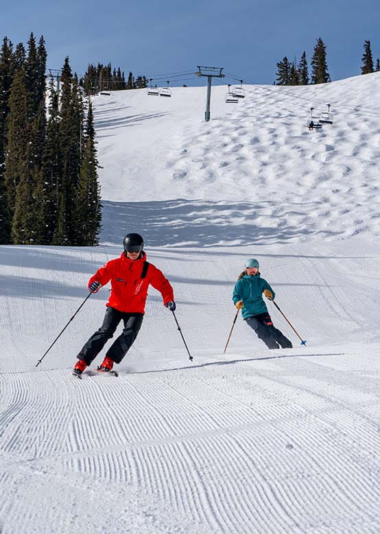 Two skiers glide down a slope, one in red, the other in green, with chairlifts in the background.