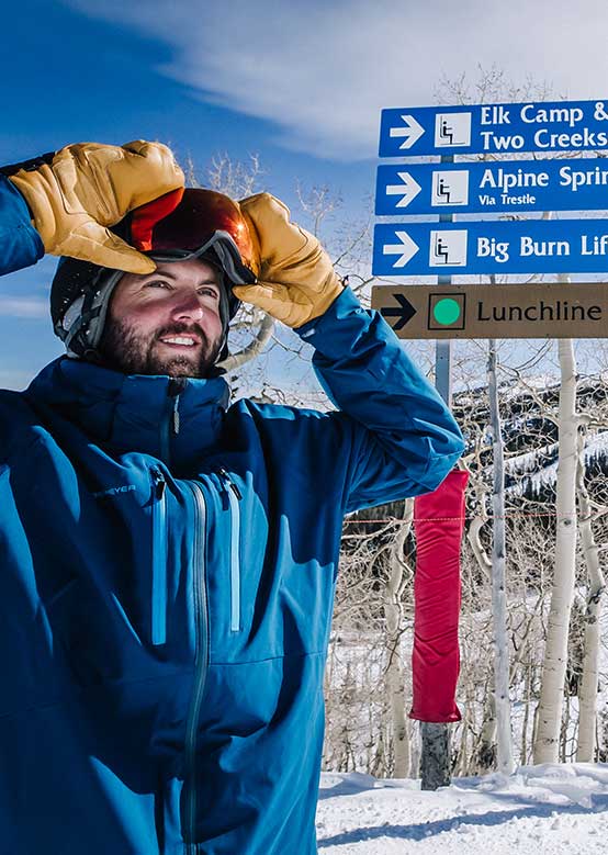 A skier adjusts goggles by snow-covered trees with signs for Elk Camp, Big Burn Lift, and Lunchline.