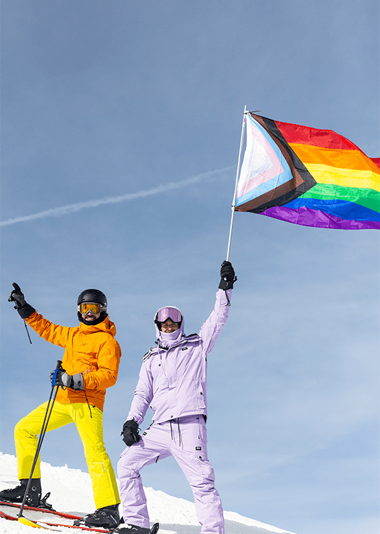 Two skiers stand proudly on snow, one in orange, the other in lavender, holding a pride flag.