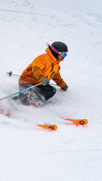 Person in orange ski outfit and matching skis carving through powder snow
