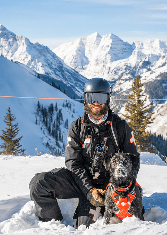 Aspen Snowmass patroller posses with his patrol dog on top of the mountain, the dogs face and snout is covered in snow