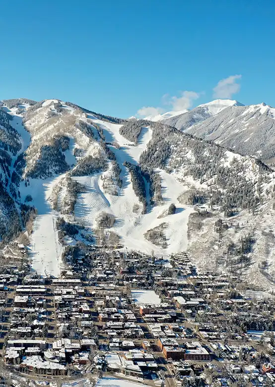 Snow-covered ski slopes descend from rugged mountains, overlooking a town under a clear blue sky.