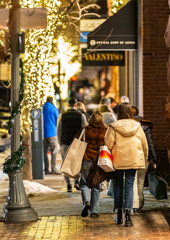 Shoppers stroll a festive, lit street with bags, snow patches lining the sidewalk amid holiday decorations.