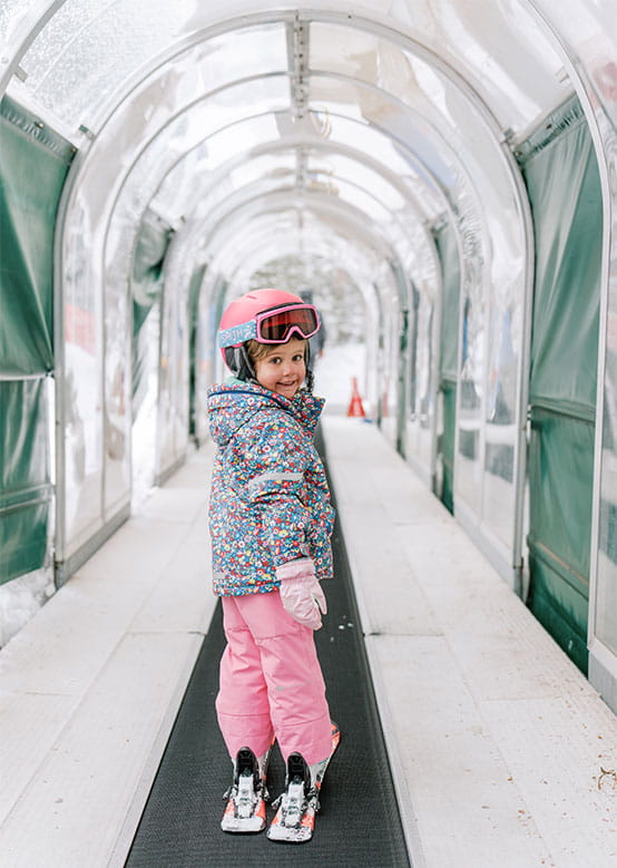 A child in a colorful snowsuit and pink helmet smiles on skis in a snow-lined arched bunny hill.