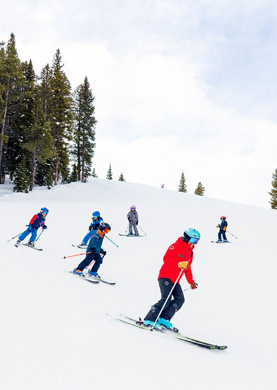 Skiers descend a snowy slope, led by a ski instructor in red, with colorful gear and evergreen trees nearby.