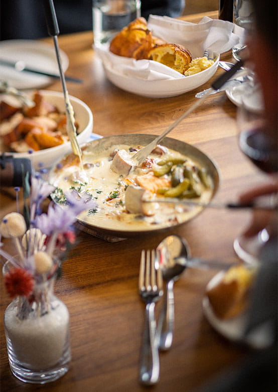 A creamy dish is served from a bowl at a table with other dishes and a vase of dried flowers.