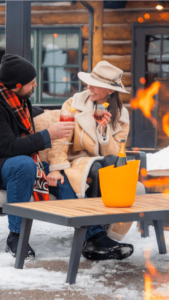 Couple sits in front of a fire outdoors at a snowcat dinner as they enjoy their cocktails at the Cabin