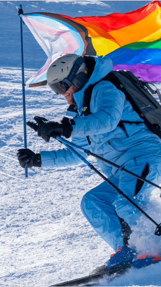 Person in blue ski pants and jacket skies down aspen highlands with pride flag for aspen gay ski week