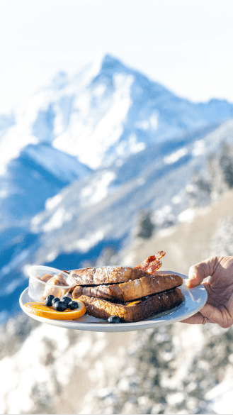 plate of french toast made by the buttermilk cliff house, held in front of the maroon bells from the cliffhouse lookout