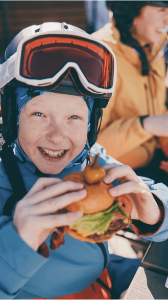 Little boy smiles as he takes a bite out of a huge cheeseburger with his ski gear on 
