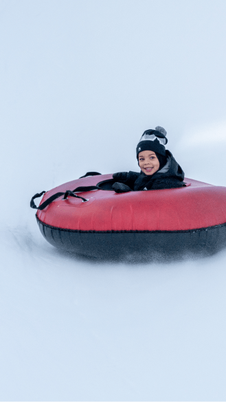 Little boy smiles as he wizzes down a white tubing hill in a red tube, at snowmass