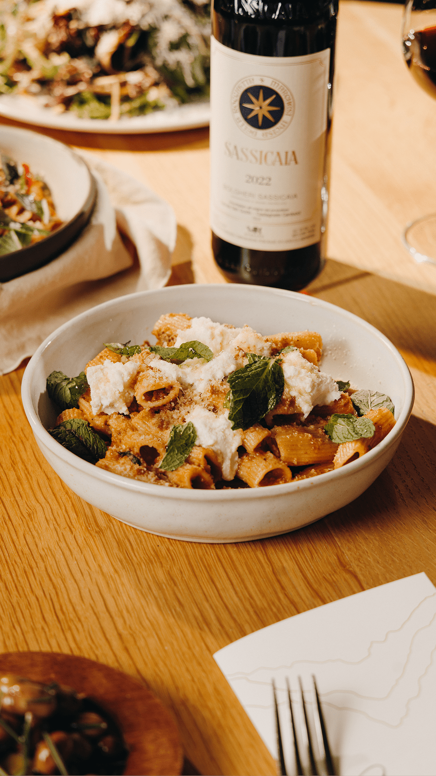 Bowl of homemade pasta with bottle of red behind, with various other plates on the table