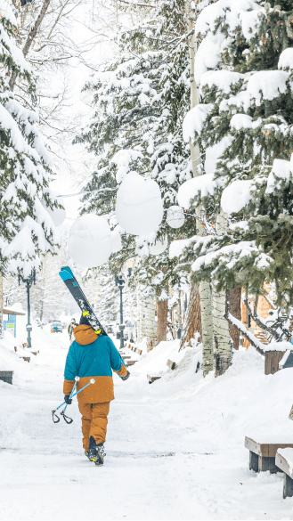 man walks down a snow packed walking mall in Aspen, with his skies over his shoulder, no one else is in sight
