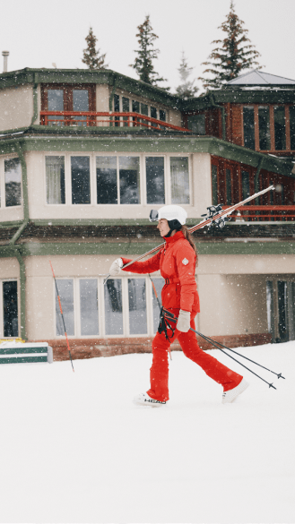 woman dressed in a red Aspen Collection snow suit walks past the little nell on a winter day, her skies over her shoulder