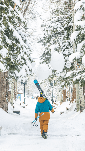 man walks through town of aspen while snow falls, skies on his shoulder