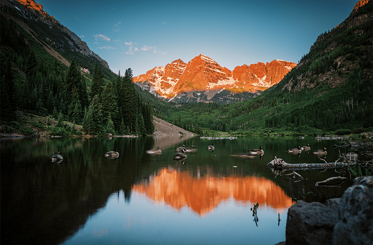 Sunrise at the maroon bells, sunlit mountains in background, ducks swim in lake in foreground 