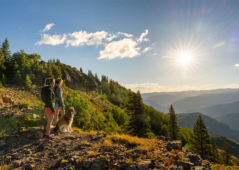 Two people and their dog stand on a lookout at Aspen Snowmass, at the top of their hike