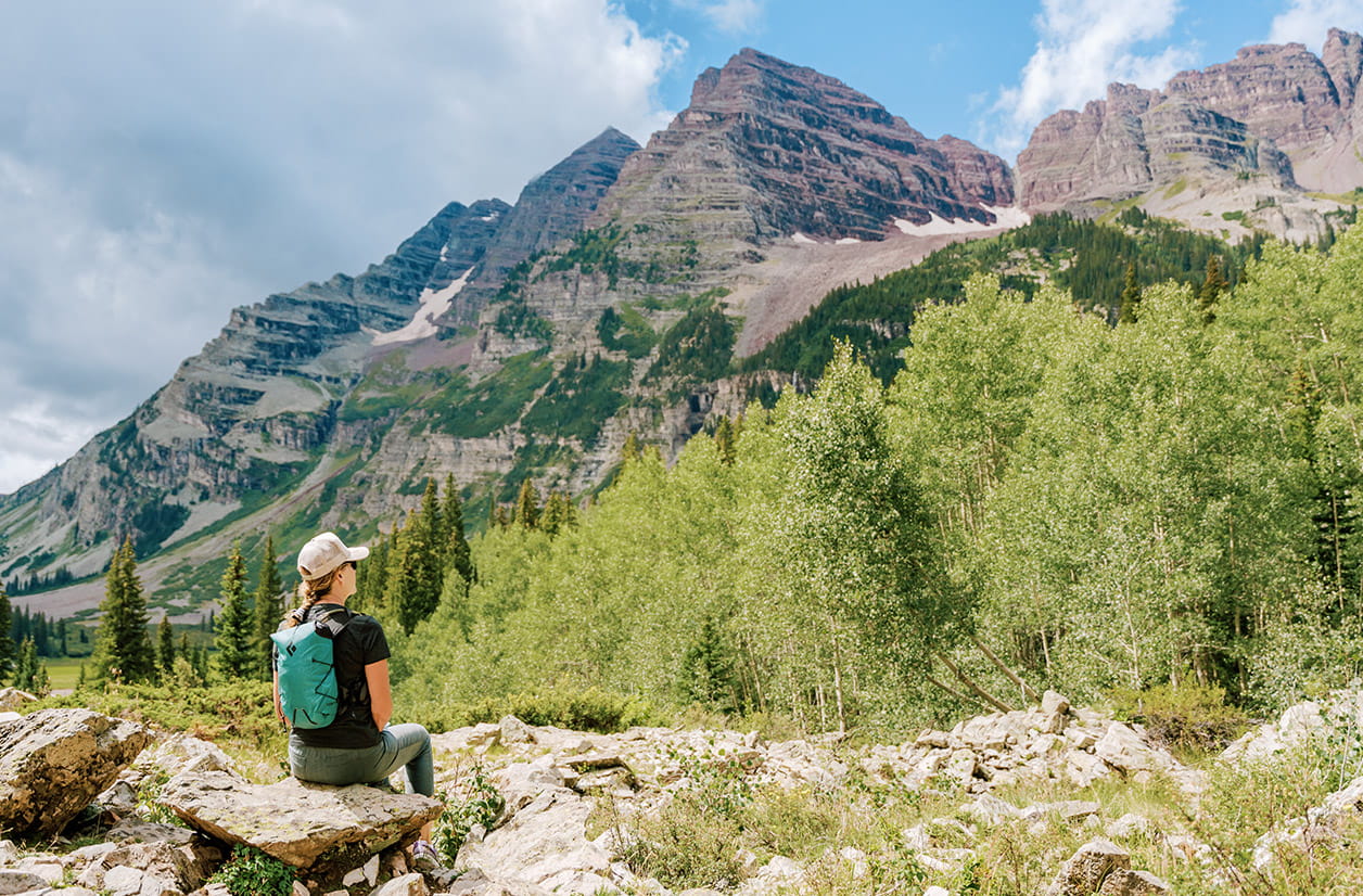 Woman in blue backpack sits on a rock on the Maroon Wilderness hiking trail, she takes break as she takes in the huge Maroon Bells in front of her, dark clouds start to gather on the horizon