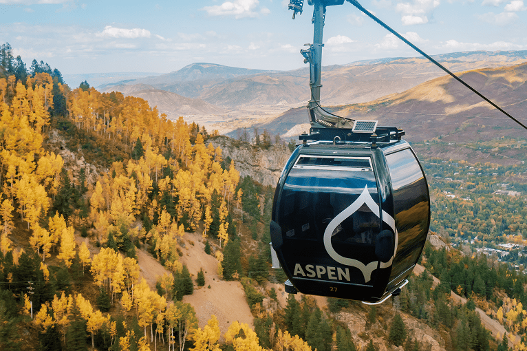 single black gondola in the forefront, as is travels up aspen mountain, with bright orange trees behind and the roaring fork valley below