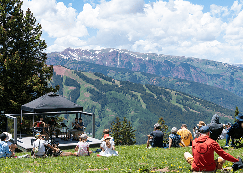 Dispersed crowd sits in the grass atop aspen mountain on a sunny summer day listening to a live classical band play in a pergola 
