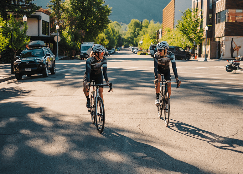 Two cyclists talk to one another as they ride down a street in Aspen on their road bikes rented from Aspen Collection