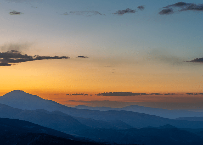 mountain range at sunset, the mountains glow dark blue with the sunset casting a glow on them, orange, yellow and dark blue sky above