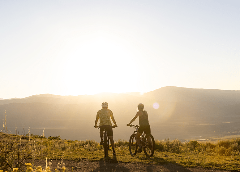 Two cyclists look out over the view atop Snowmass, as the sun sets over the mountains