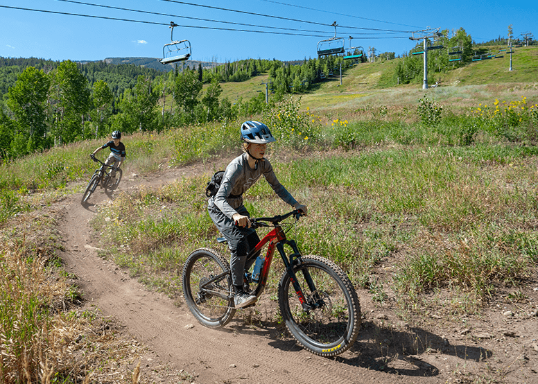 Two young kids take a turn on a single track trail on their mountain bikes, the snowmass chair lift travels above them