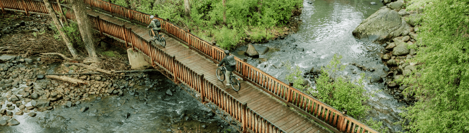 Aerial view of a group ebiking across a wooden bridge over the river surrounded by green trees in Aspen Colorado