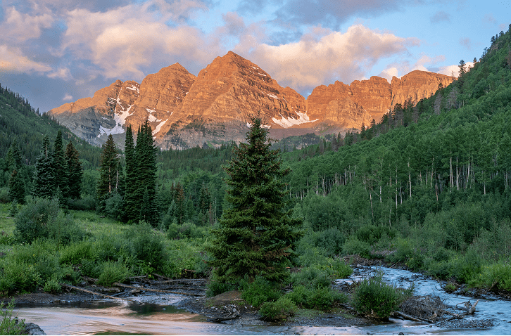 Maroon Bells under sunny summer skies, the sun setting casts a golden glow on the peaks