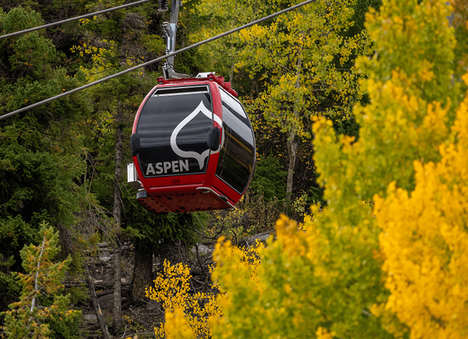Single red gondola going up aspen mountain, in the green and yellow aspen trees during a cloudy summer day