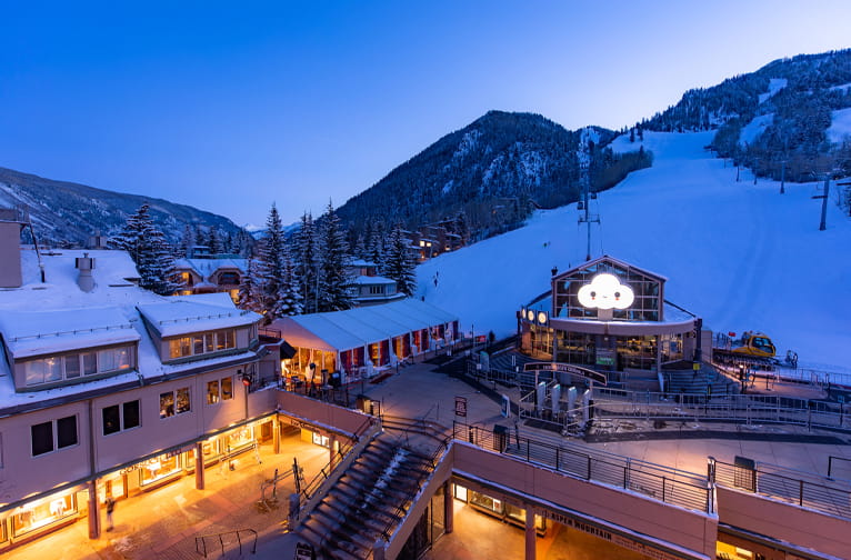 Ski resort buildings glow under evening light, nestled at the base of a snowy mountain with slopes.