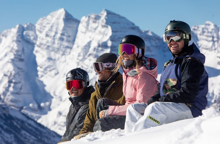 Four friends in Aspen winter gear laugh on a snowy ledge, with towering sunlit mountains behind them.