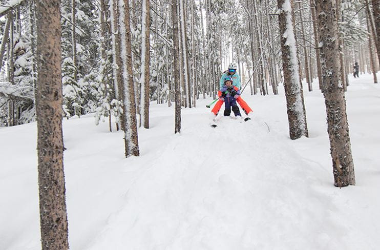 A dad shows his daughter how to ski through a tree-lined trail at Snowmass