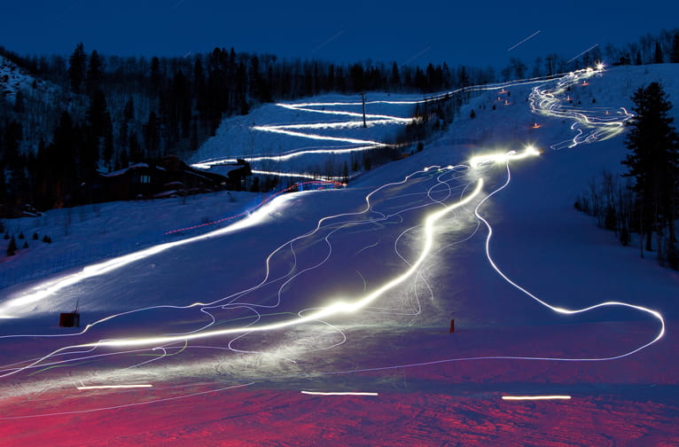 Light trails follow skiers down a snowy night slope, with dark trees and lit paths under a starry sky.