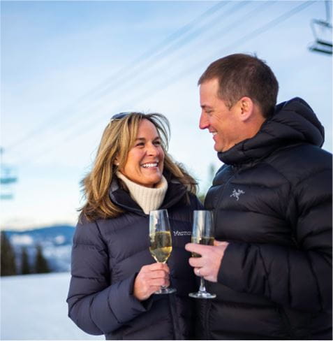 Couple smiling with glasses of champagne under a chairlift during the winter